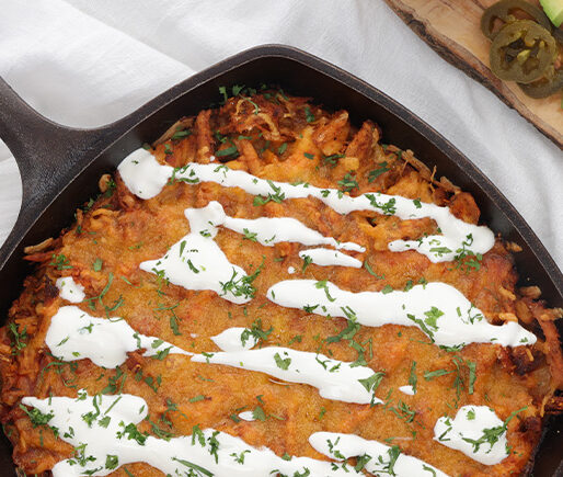 Cast-iron skillet with a cheesy baked dish drizzled with crema and sprinkled with cilantro and herbs, on a light table with avocado and lime nearby.