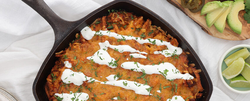 Cast-iron skillet with a cheesy baked dish drizzled with crema and sprinkled with cilantro and herbs, on a light table with avocado and lime nearby.
