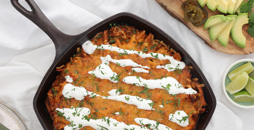 Cast-iron skillet with a cheesy baked dish drizzled with crema and sprinkled with cilantro and herbs, on a light table with avocado and lime nearby.
