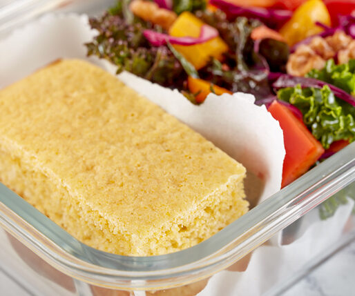 A slice of cornbread rests in a clear lunchbox beside a colorful mixed-garden salad with greens and vegetables.