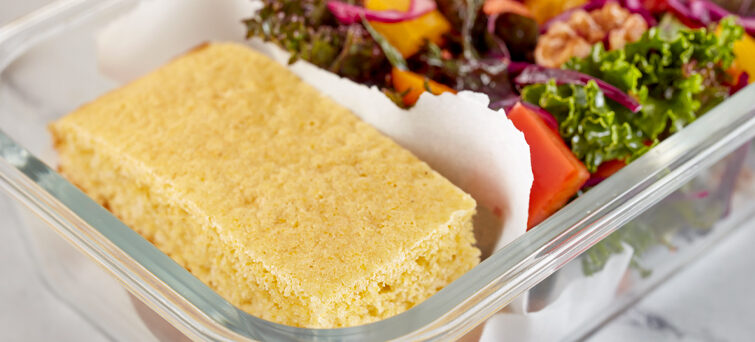 A slice of cornbread rests in a clear lunchbox beside a colorful mixed-garden salad with greens and vegetables.