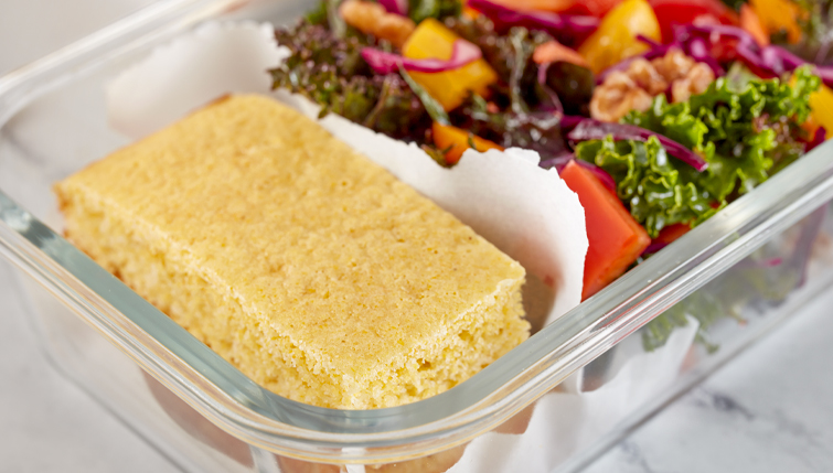 A slice of cornbread rests in a clear lunchbox beside a colorful mixed-garden salad with greens and vegetables.