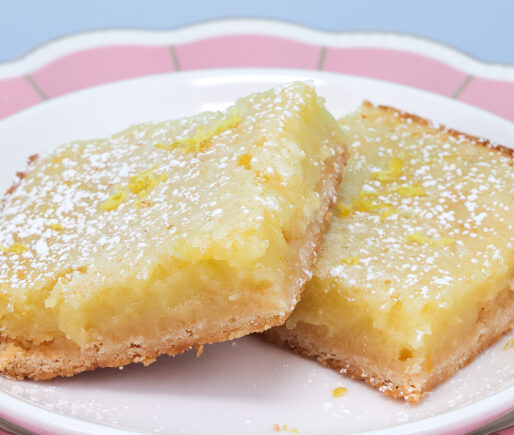 Two lemon bars with powdered sugar on a white plate with a pink rim against a blue background.