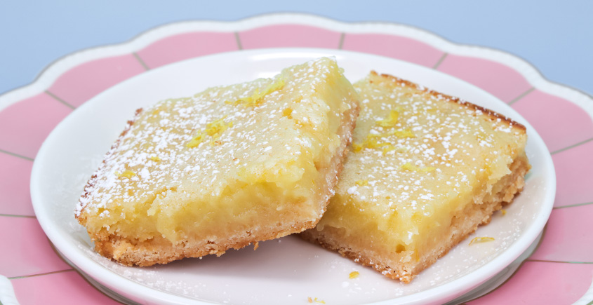 Two lemon bars with powdered sugar on a white plate with a pink rim against a blue background.