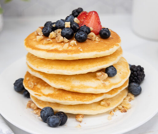 Stack of four fluffy pancakes topped with blueberries and a strawberry, sprinkled with chopped nuts, on a white plate.