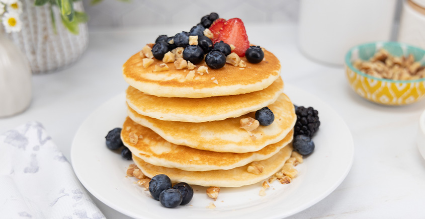 Stack of four fluffy pancakes topped with blueberries and a strawberry, sprinkled with chopped nuts, on a white plate.