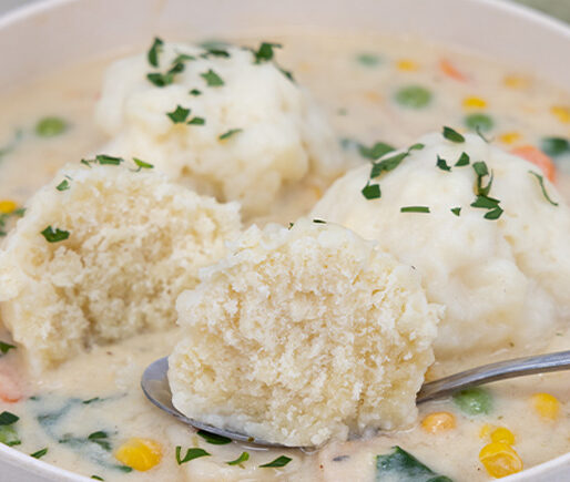 Creamy chicken and dumplings soup in a white bowl with corn, peas, and carrots, garnished with parsley; a spoon holds a dumpling.