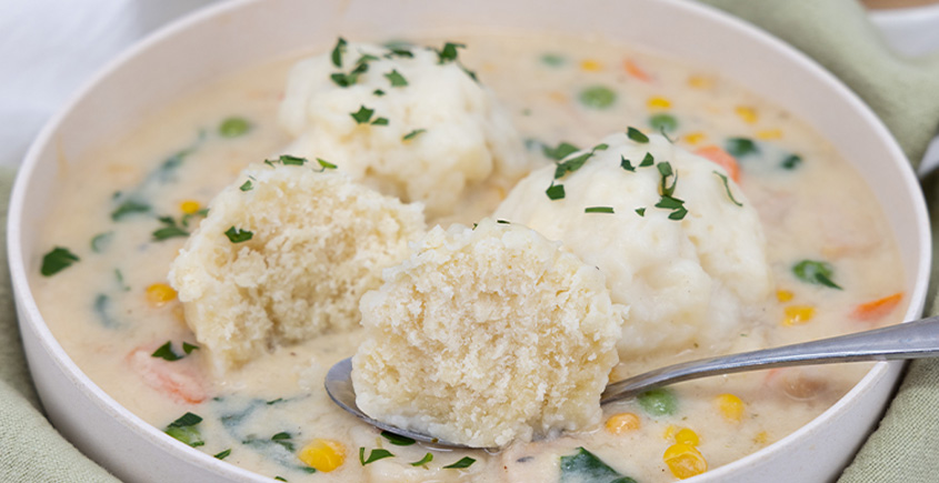 Creamy chicken and dumplings soup in a white bowl with corn, peas, and carrots, garnished with parsley; a spoon holds a dumpling.