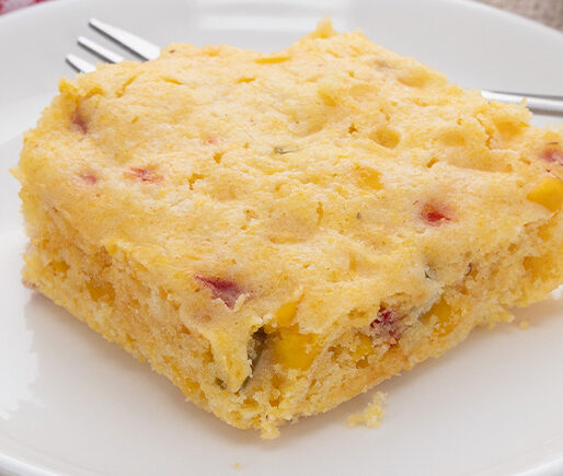 Slice of cornbread with corn kernels and red pepper on a white plate with a fork nearby