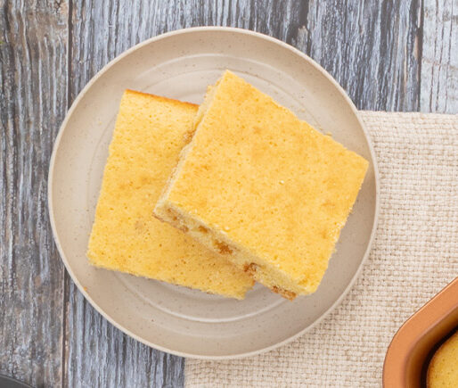 Two square vanilla sponge cake pieces on a beige plate atop a rustic wood table, with part of a baking pan visible nearby.