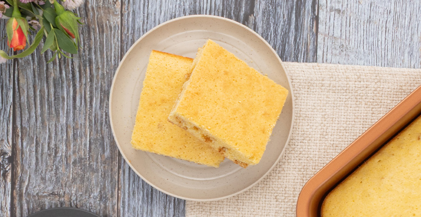 Two square vanilla sponge cake pieces on a beige plate atop a rustic wood table, with part of a baking pan visible nearby.