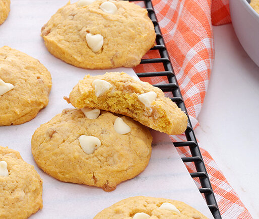 Golden cookies with white chocolate heart chips cooling on parchment on a wire rack, with an orange checkered towel nearby.