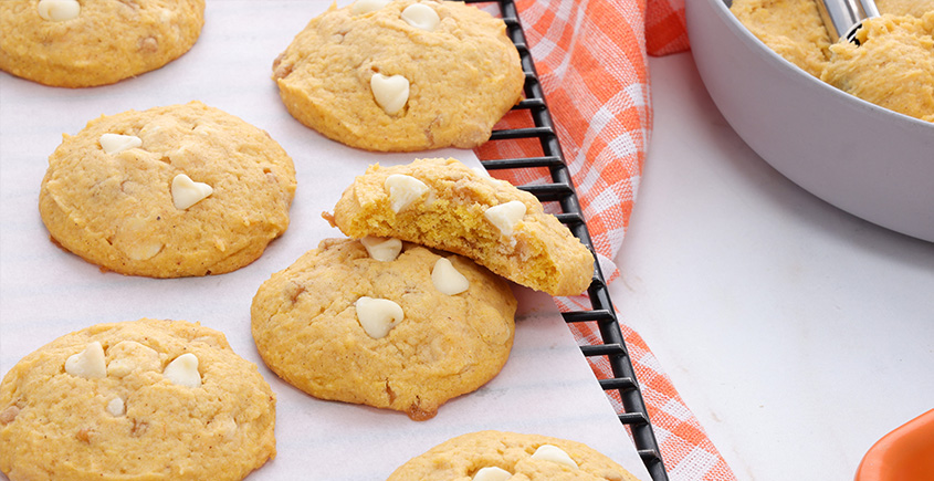 Golden cookies with white chocolate heart chips cooling on parchment on a wire rack, with an orange checkered towel nearby.