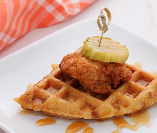 Crispy fried chicken atop a waffle wedge, topped with a pickle slice on a skewer, served on a white plate with a napkin nearby.