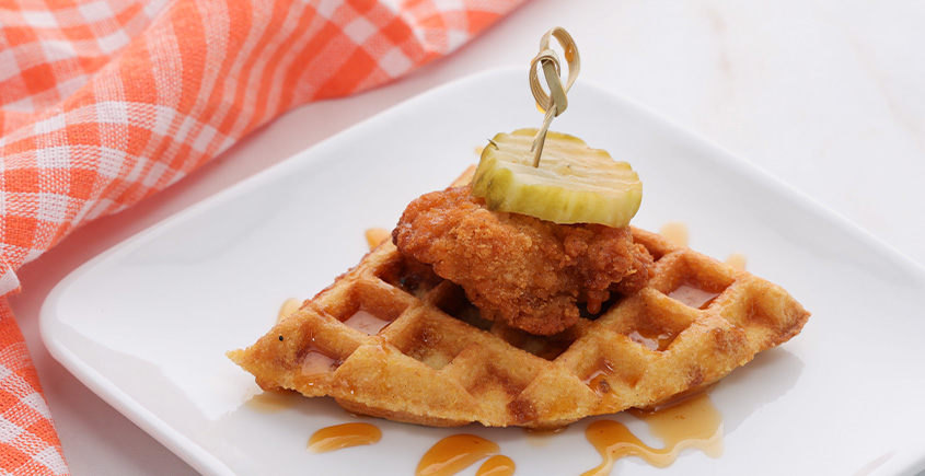 Crispy fried chicken atop a waffle wedge, topped with a pickle slice on a skewer, served on a white plate with a napkin nearby.