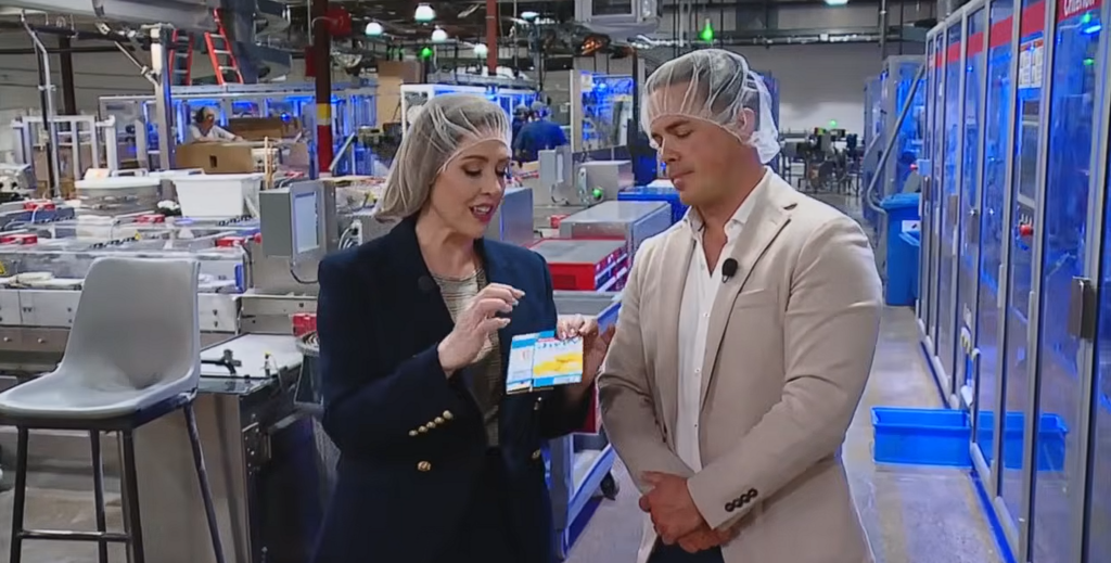 Two professionals wearing hairnets in a factory discuss a product sample card near an automated production line.