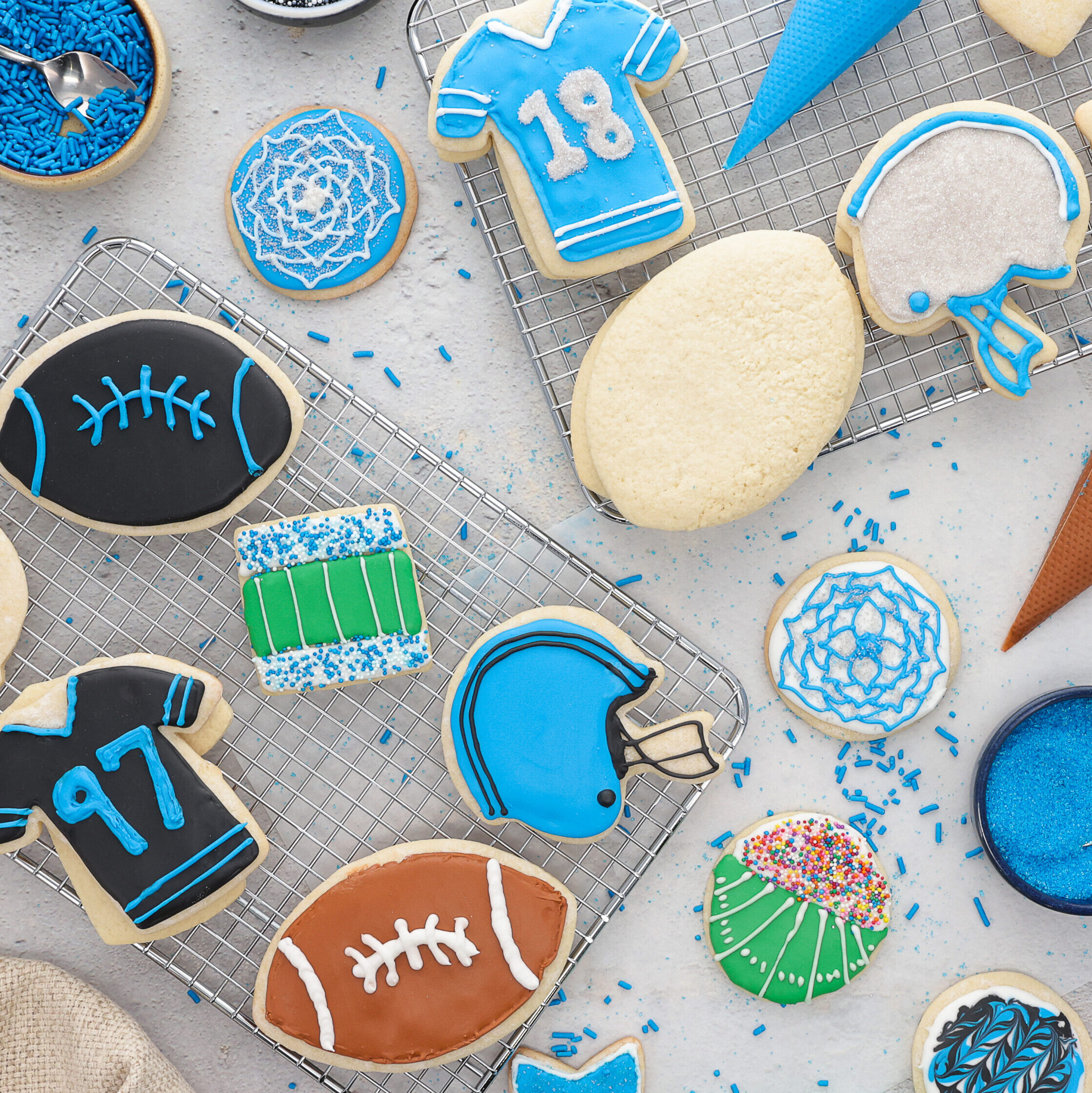 Assorted blue and white sports-themed cookies on a cooling rack, including a football, jersey with the number 18, and helmet shapes.