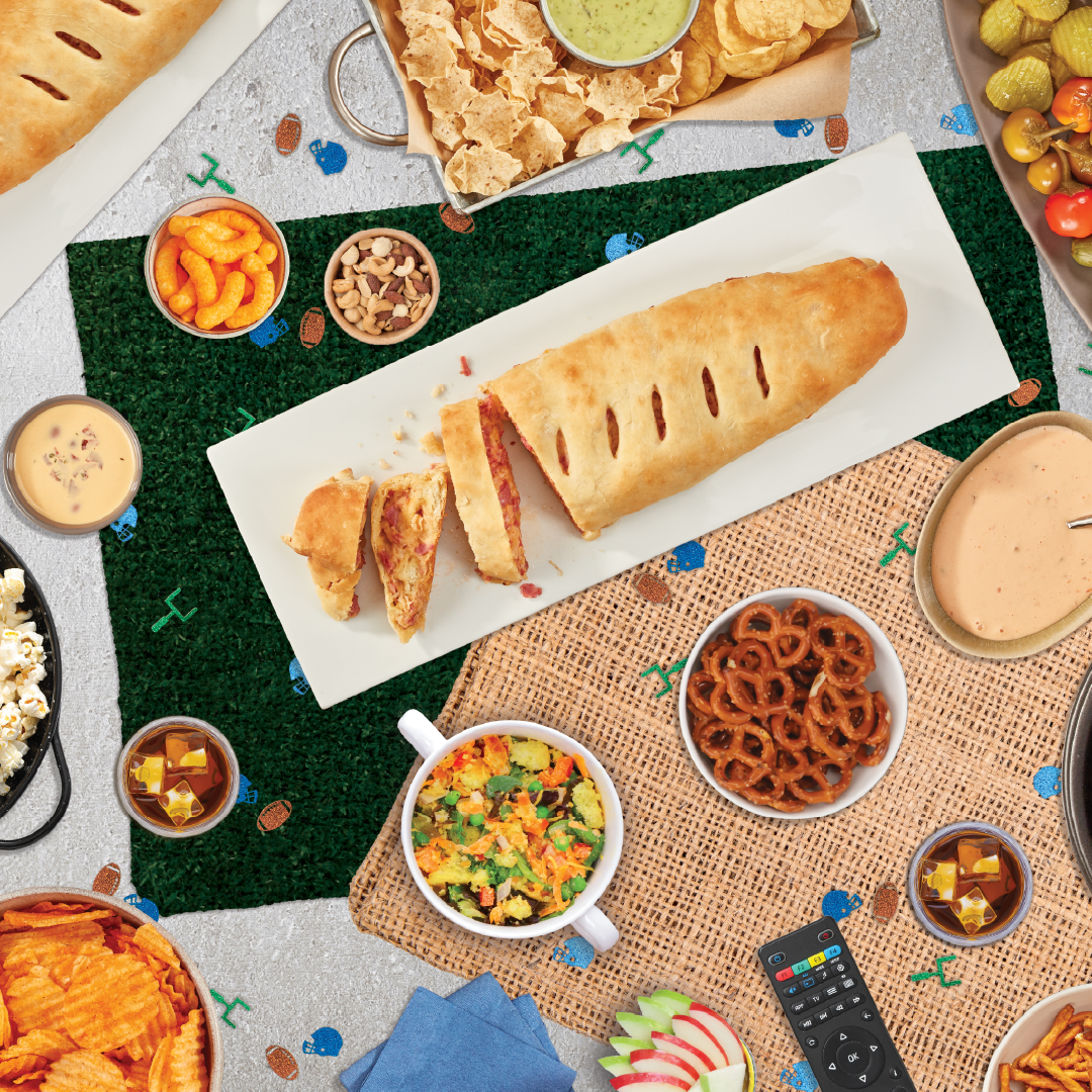 Top-down view of a party spread on a turf placemat: a sliced baguette on a white board, chips, nuts, pretzels, dips, and drinks.