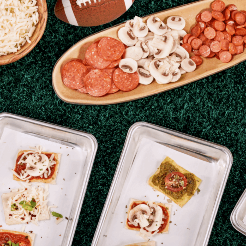 Snack spread on artificial turf featuring pepperoni and mushroom toppings on a wooden tray and small pizzas on metal trays with a football nearby.