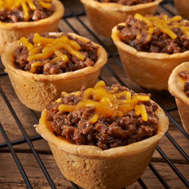 Mini taco cups filled with seasoned ground beef and melted cheddar on a cooling rack at a party.