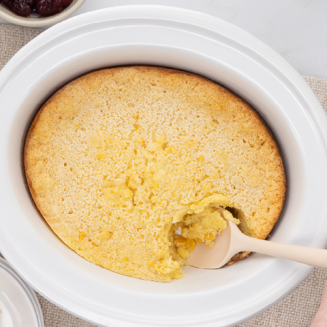 Sponge cake in a white round dish with a spoon scooping a bite from the edge, cherries in a small bowl visible at top left (table setting).